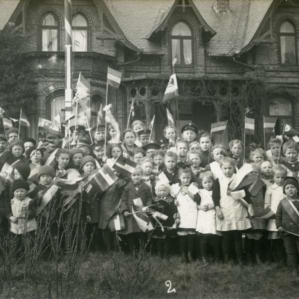 Stor forsamling af børn med det tyske flag i hånden foran en bygning i Kobbermølle. Nr. fire fra venstre i første række har to små Dannebrogssplitflag på brystet.